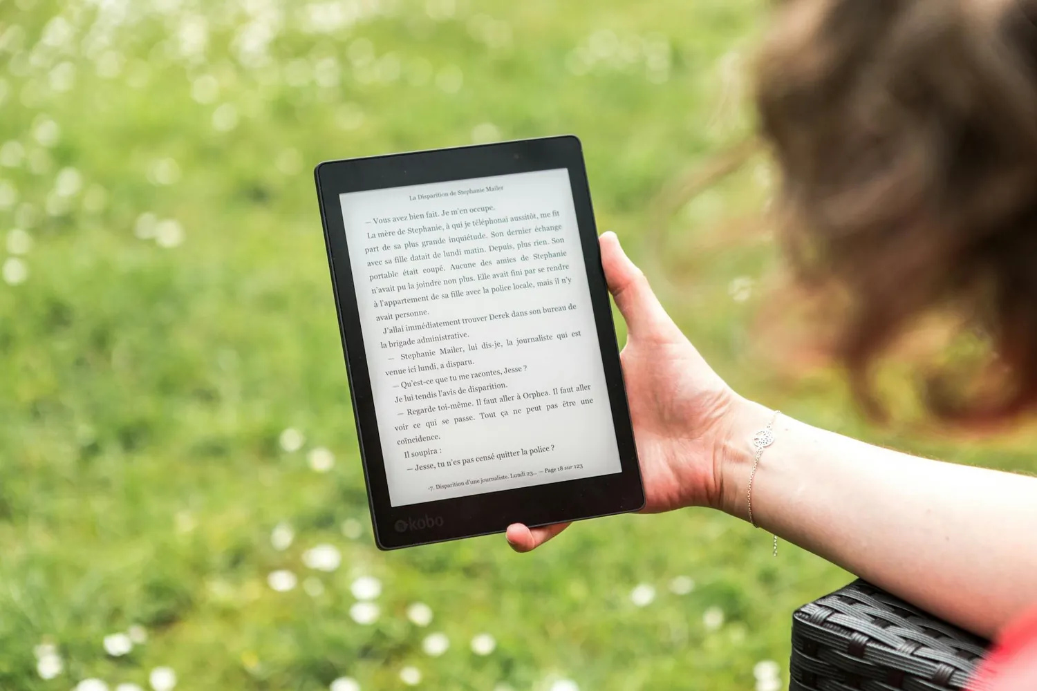A hand holding an e-reader outdoors, an open page of text on its screen against grass
