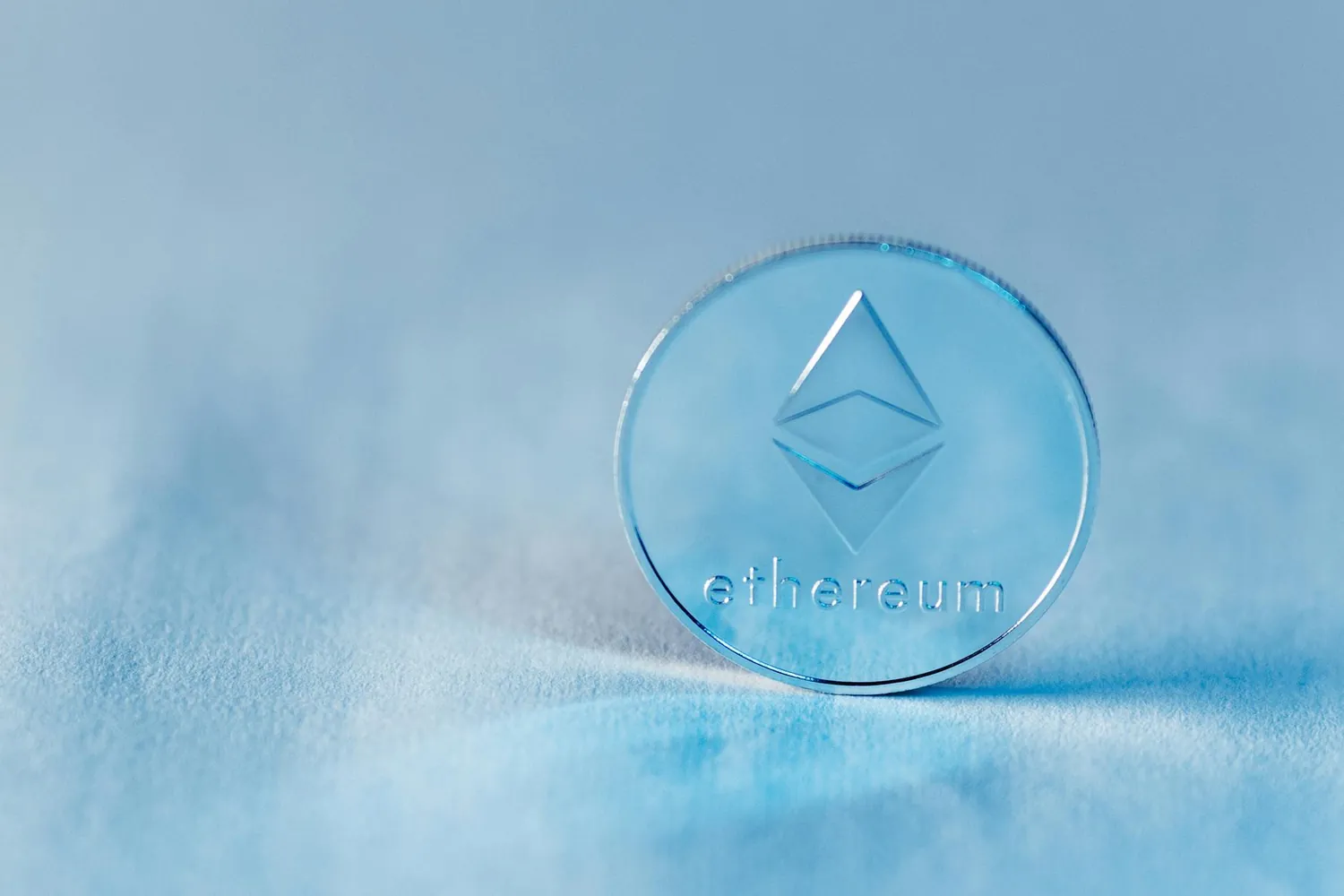 A physical coin embossed with the Ethereum symbol resting on a pale blue surface