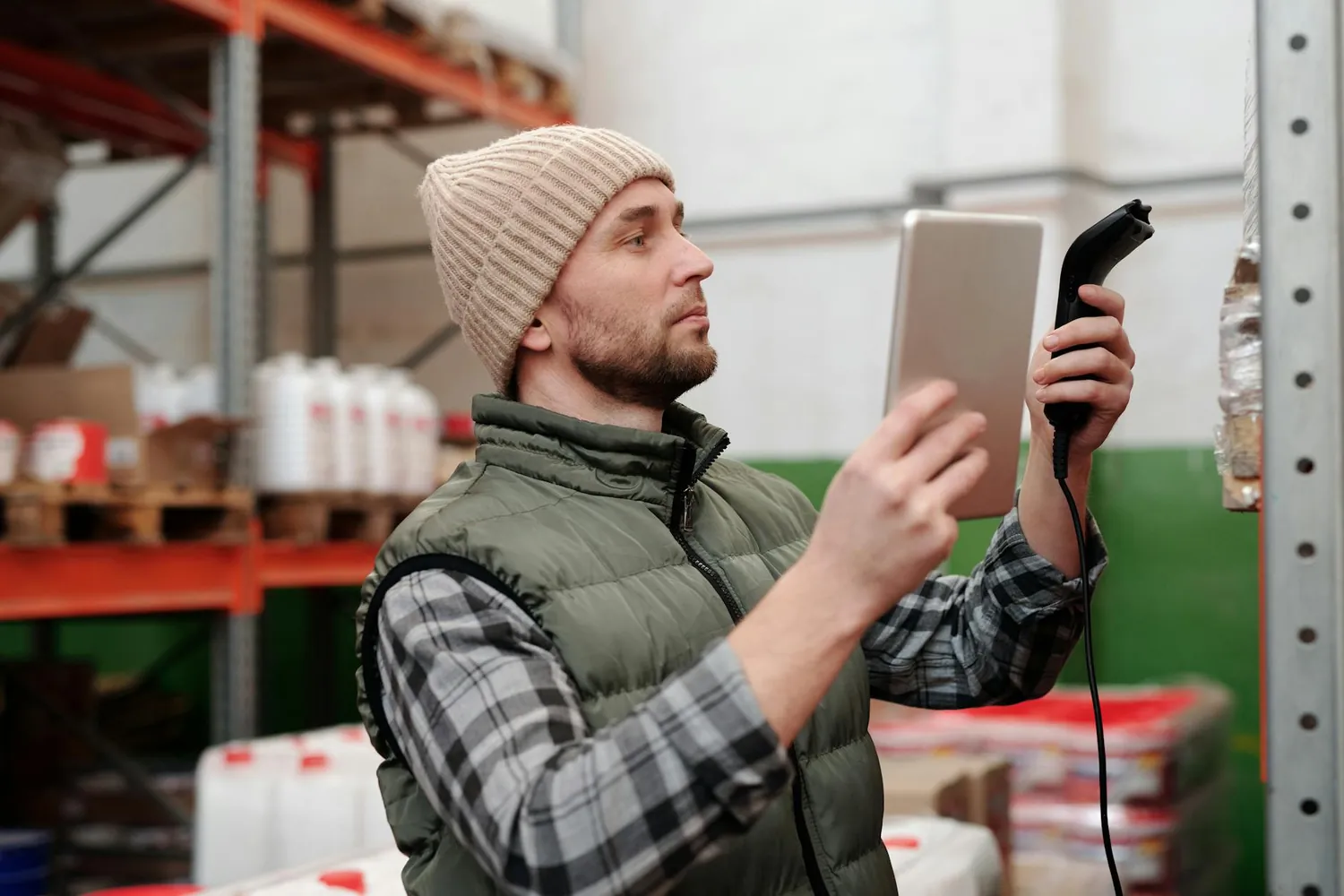 A warehouse worker in a beanie holding a tablet and a barcode scanner beside shelves of stock