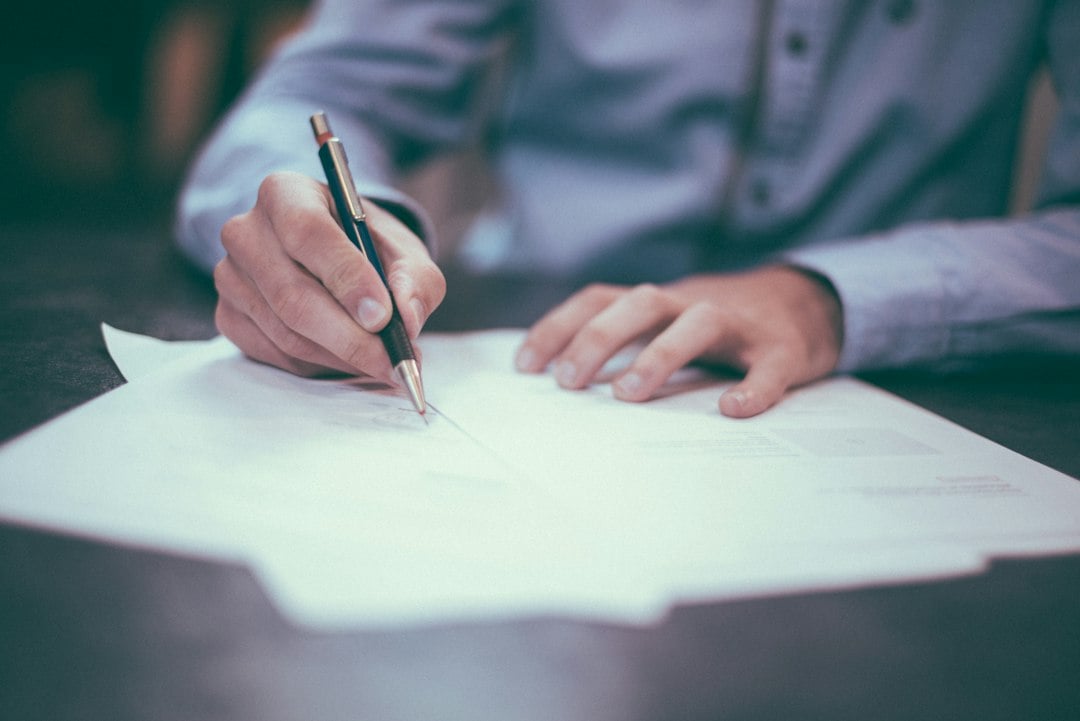 A man signs a paper document with a pen at a dark table