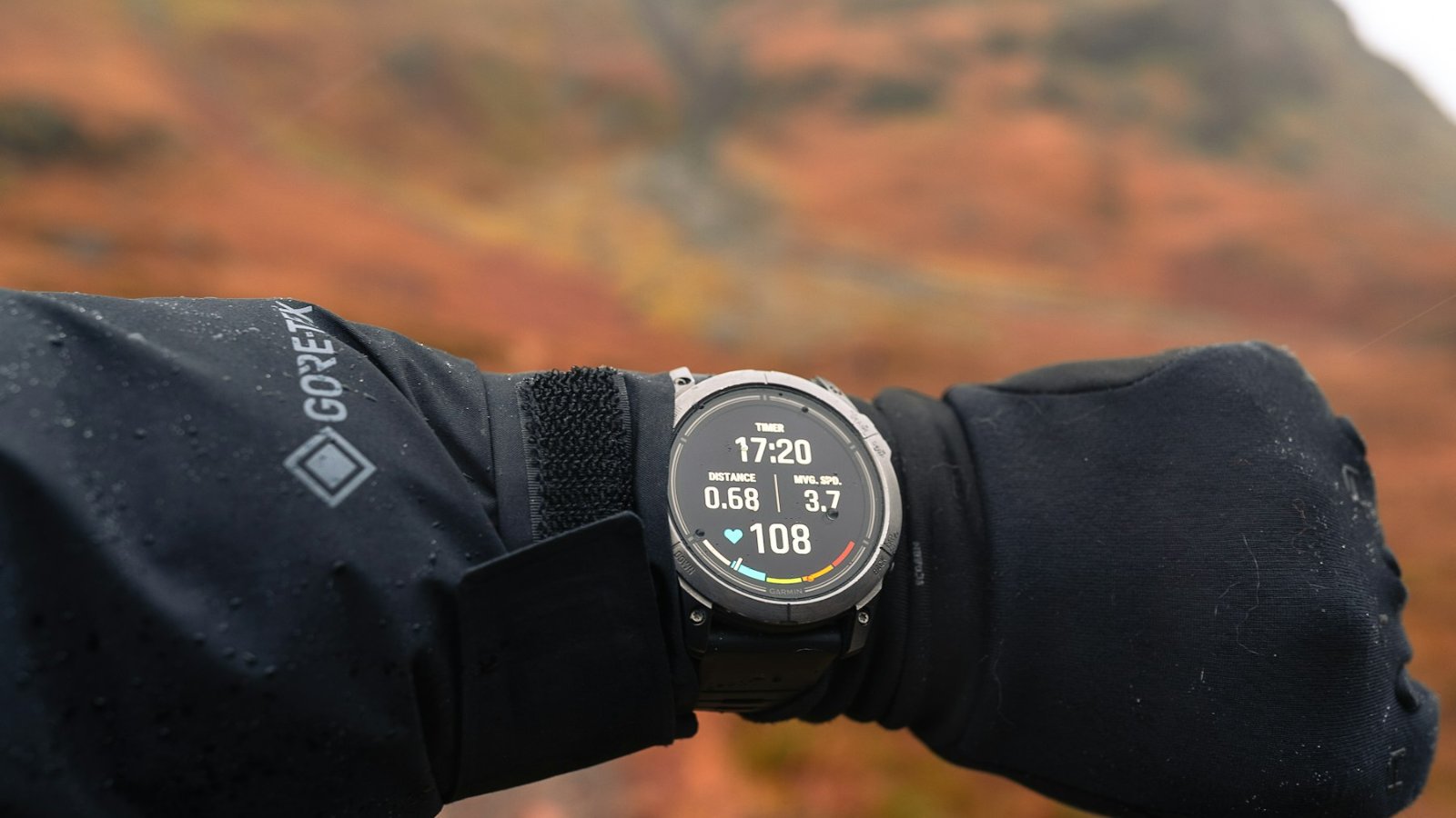 A round multisport GPS watch worn on the wrist, its memory-in-pixel screen showing elapsed time, distance, pace and heart rate, on a hillside in daylight