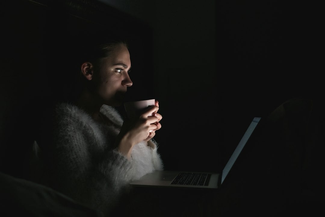 A woman holding a mug while working on a laptop in a dark room lit by the screen