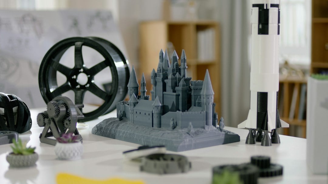 A highly detailed grey 3D printed castle on a desk beside a rocket model and filament spool