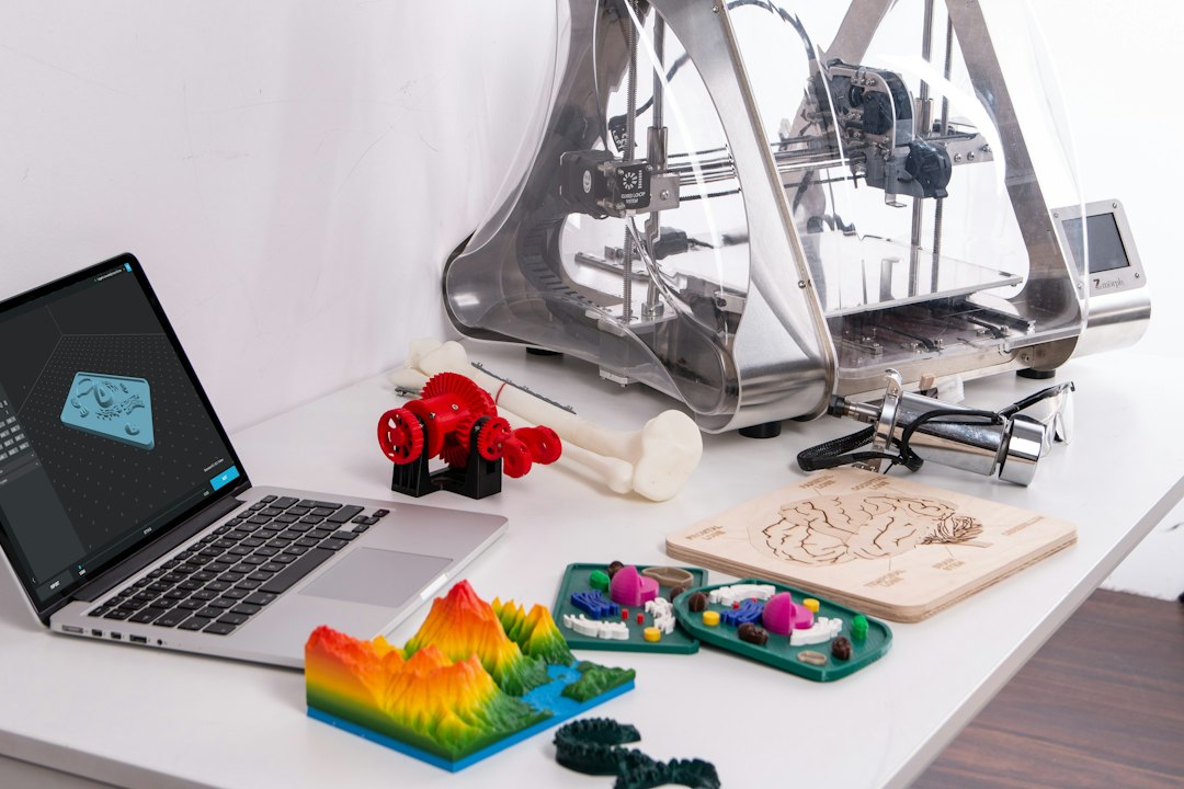 A laptop running 3D design software beside a 3D printer and an assortment of colourful printed models on a white desk