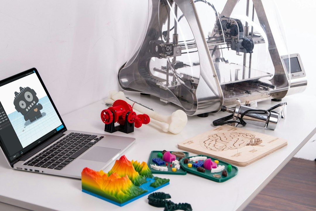 Laptop showing a 3D robot model next to a 3D printer and printed figurines on a white desk