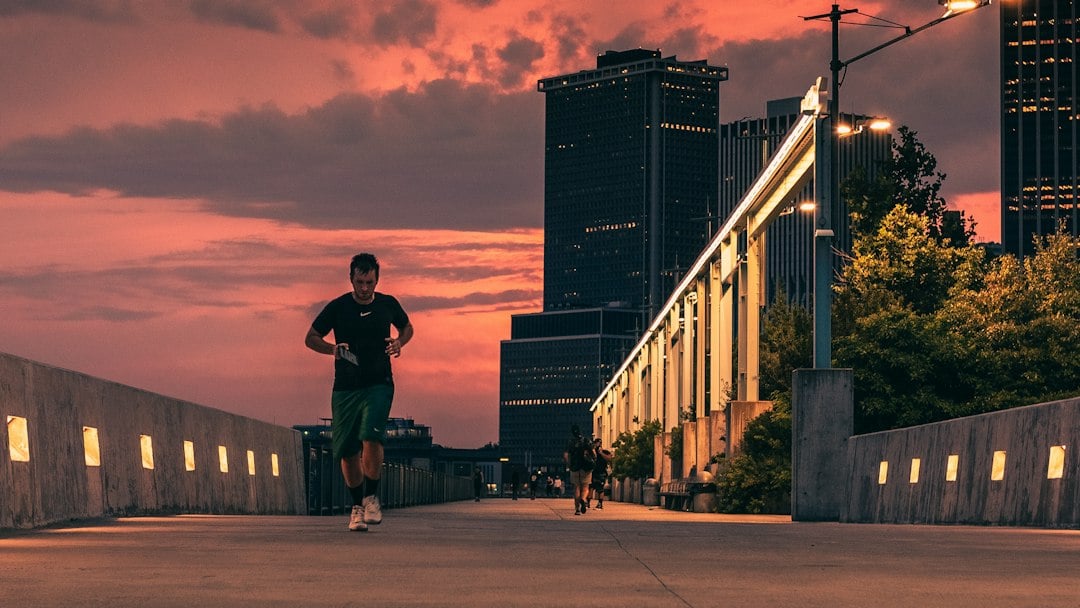 A runner in dark training kit runs along a raised walkway at sunset with city towers behind.